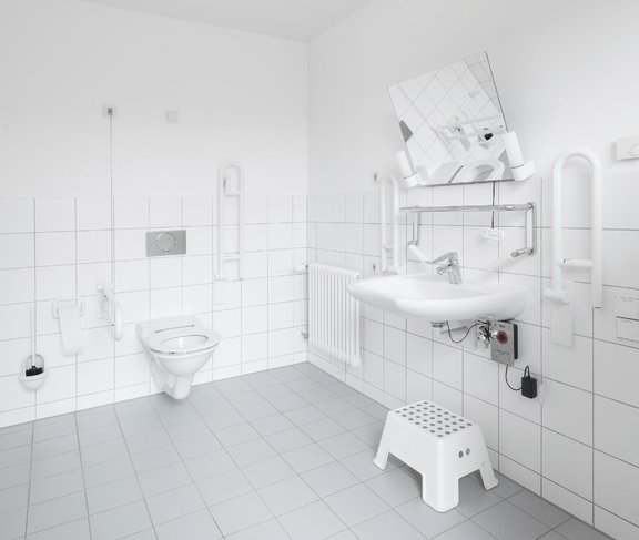 Blick auf eine behindertengerechte Toilette mit Haltegriffen, Notfallalarm und einem Hocker unter dem Waschbecken